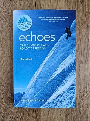 A portrait of the cover of Bullock's book 'Echoes' on a wood surface. The cover is blue showing a climber on a steep snow slope.