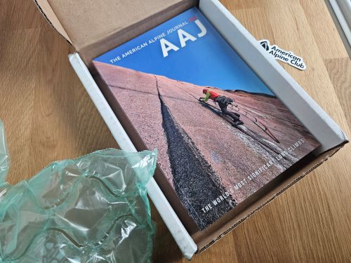 The 2025 AAJ in the shipping packaging with green bubble wrap to the left and the AAC sticker on the table to the right with the book in the middle of an aid climber on a big wall contrasting blue sky.